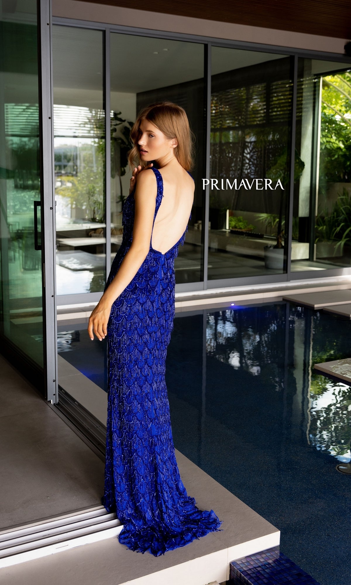 A woman in a sleeveless Beaded-Fringe Long Primavera Prom Dress 4129 with an open back stands by an indoor pool. The floor-length blue gown’s elegance is highlighted, with